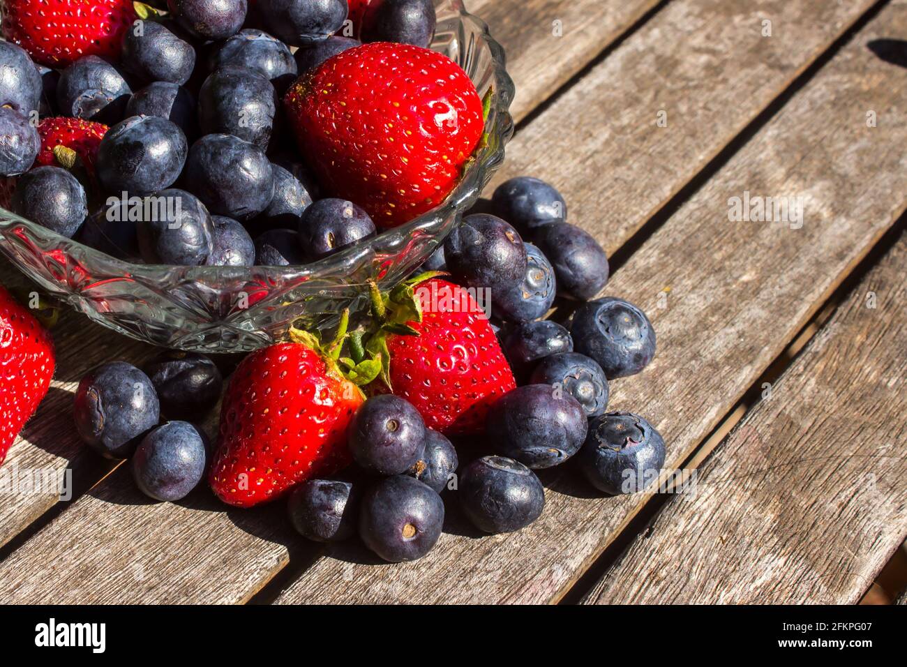A glass bowl, filled to overflowing with bright red strawberries and indigo-colored blueberries on a weathered wooden surface Stock Photo