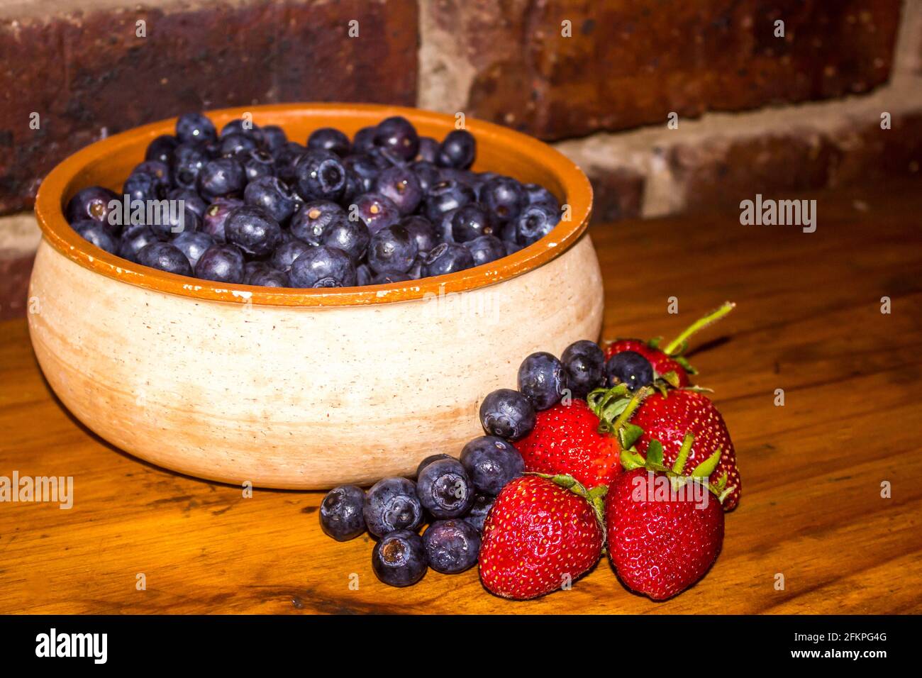 A rustic pottery bowl, filled with Indigo colored blueberries, and extra blueberries and some strawberries next to it Stock Photo