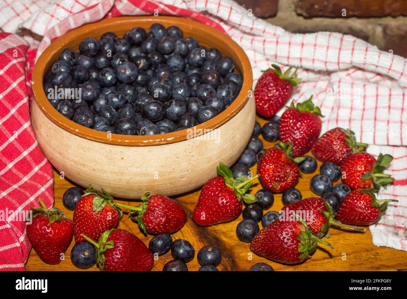 A rustic pottery bowl, filled with Indigo colored blueberries, and surrounded by more blueberries and strawberries Stock Photo