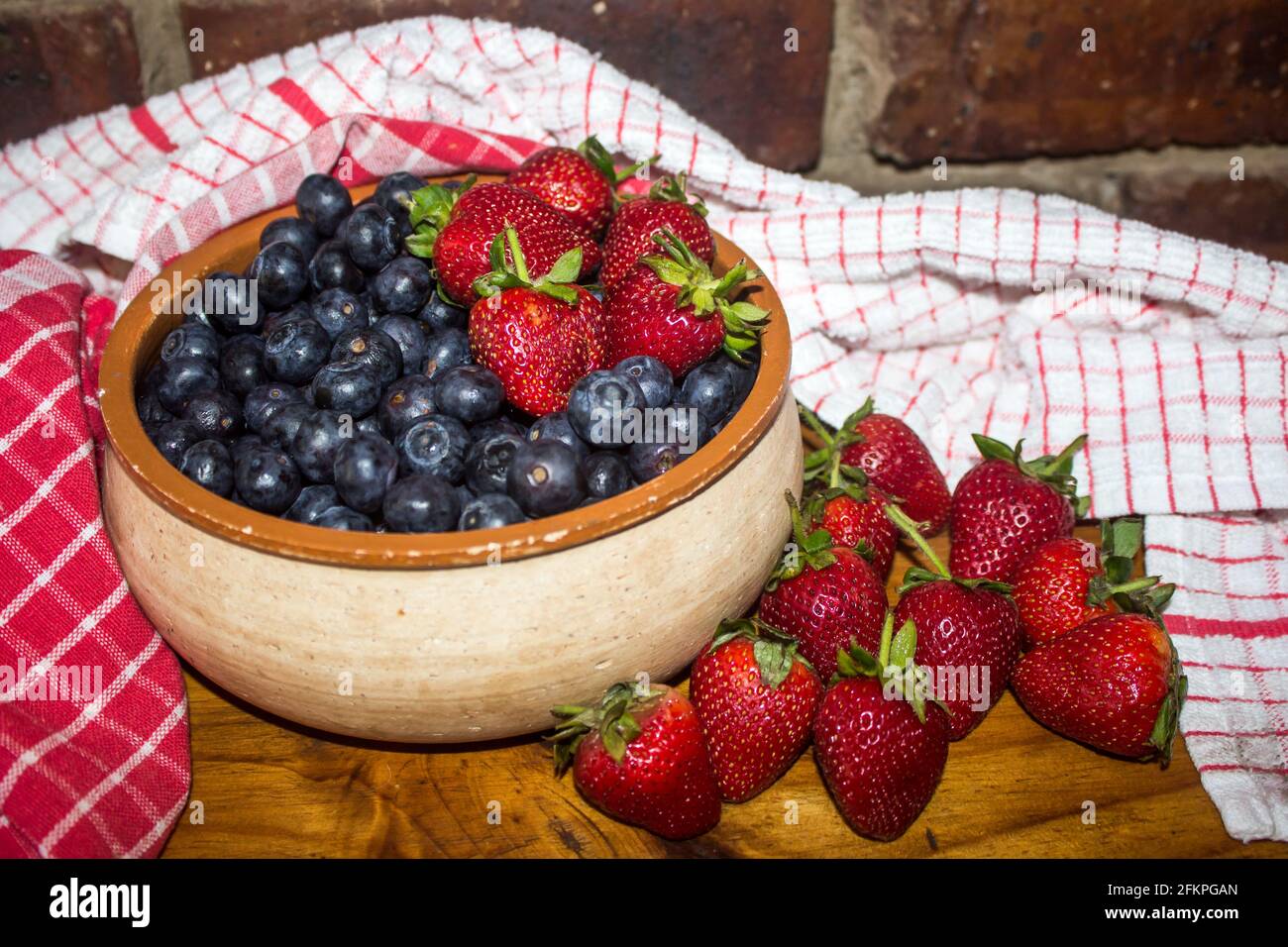 A rustic pottery bowl, filled with Blueberries, surrounded by strawberries and red kitchen Towels Stock Photo