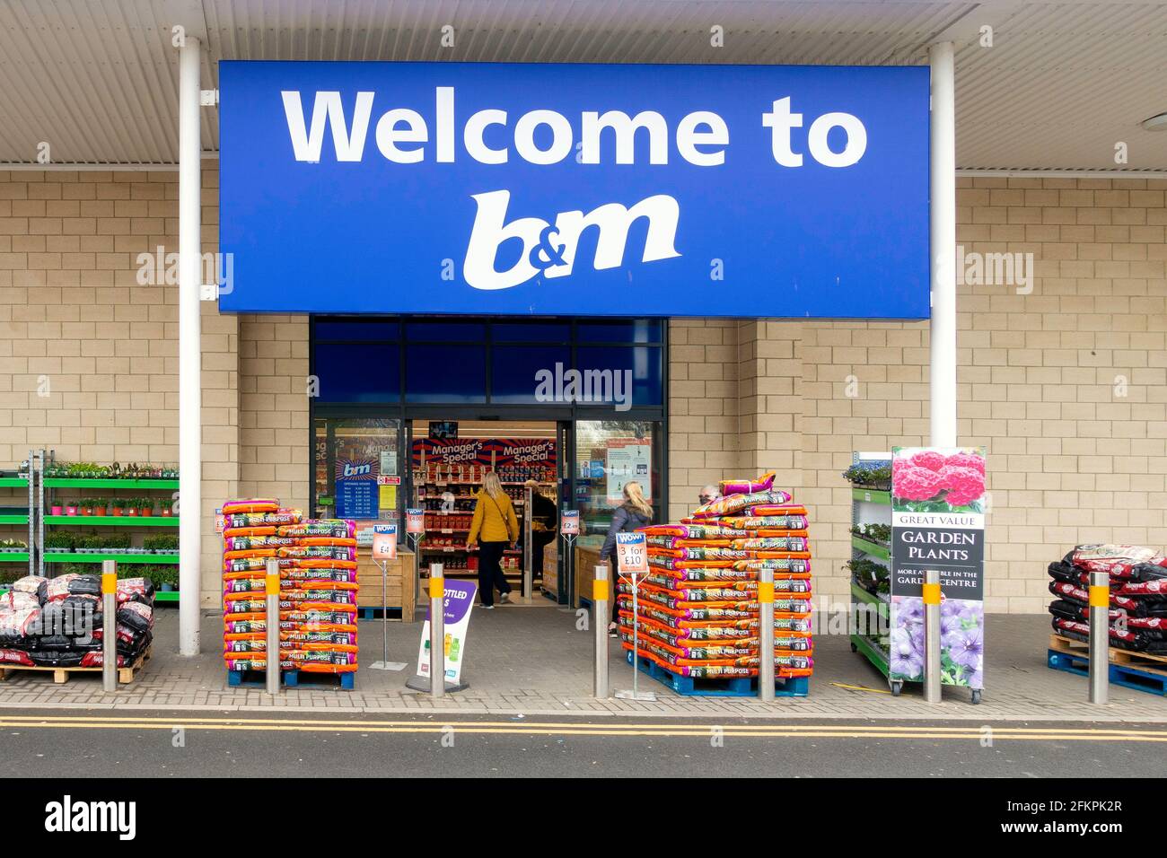 Welcome to B&M store supermarket entrance with a woman entering and piles of garden products stacked outside in Guisborough Cleveland North Yorkshire Stock Photo