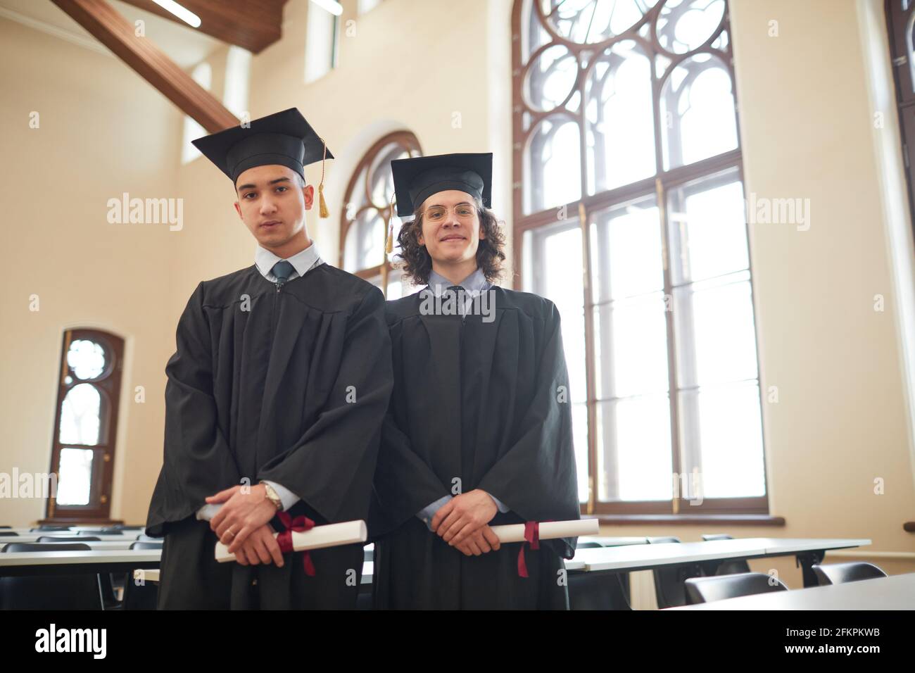 Waist up portrait of two young men wearing graduation robes and looking ...