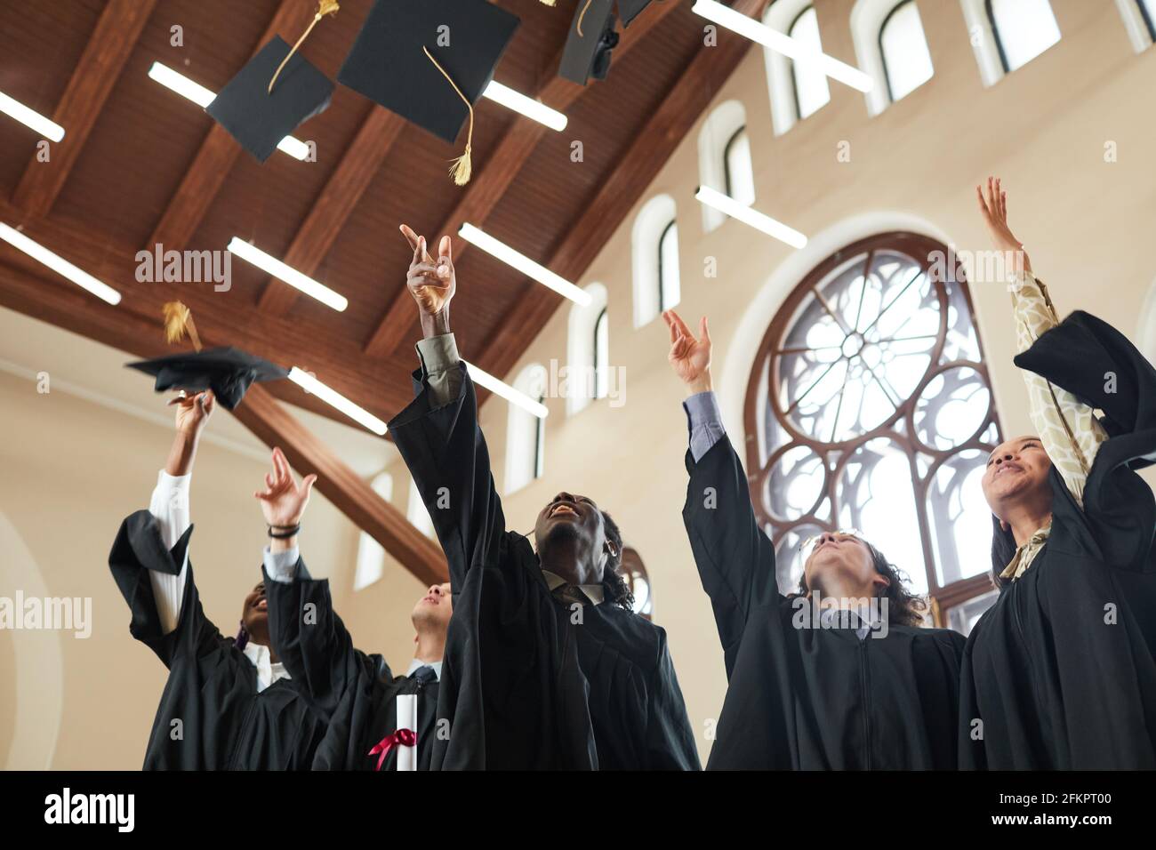 Low angle view at multi-ethnic group of young people throwing caps in air during graduation ceremony indoors in classic school auditorium, copy space Stock Photo