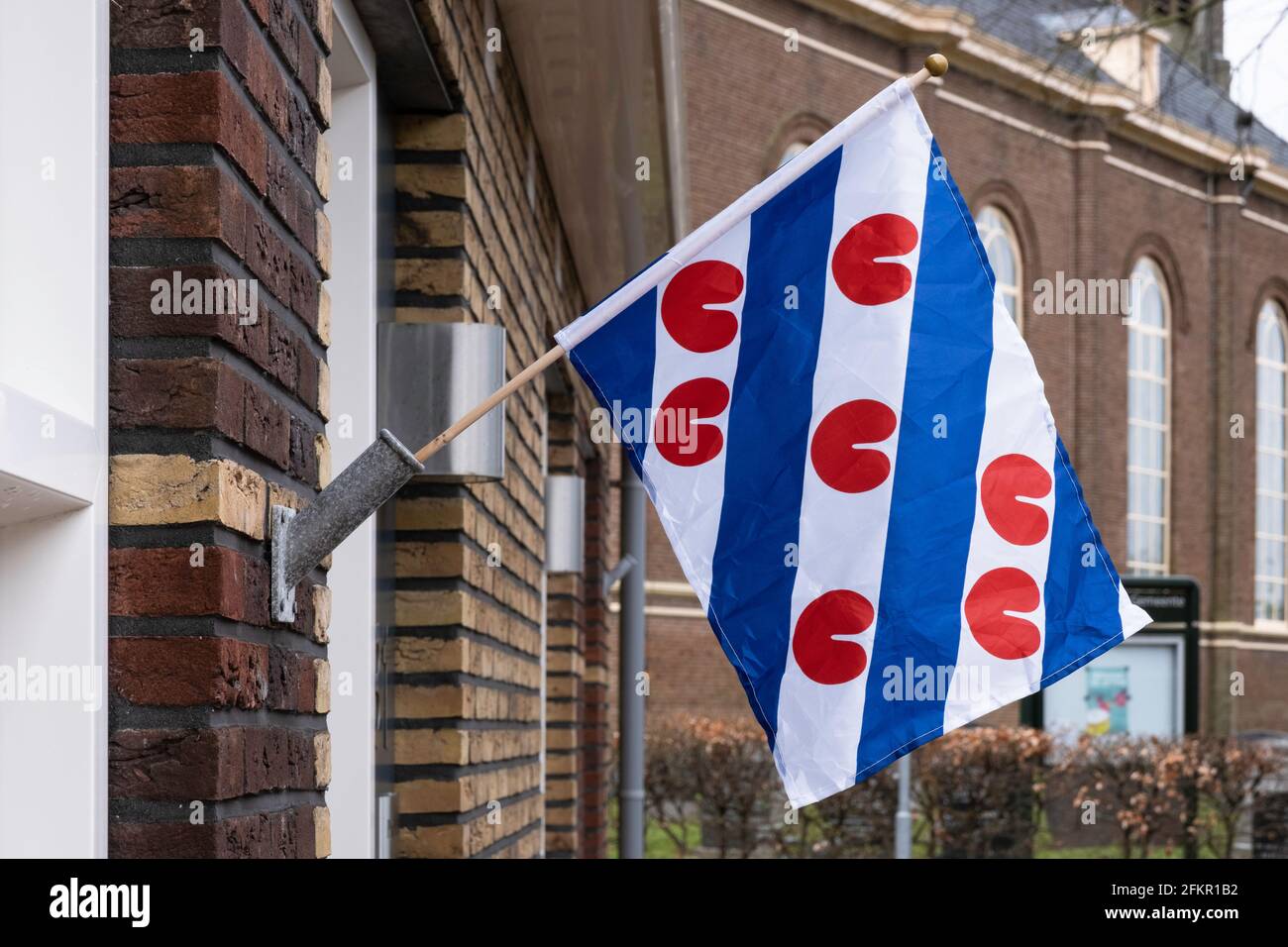Tricolor Frisian flag, provincial flag of Friesland province, hangs from a flagpole against a wall in Koudum, the Netherlands. Church in background Stock Photo