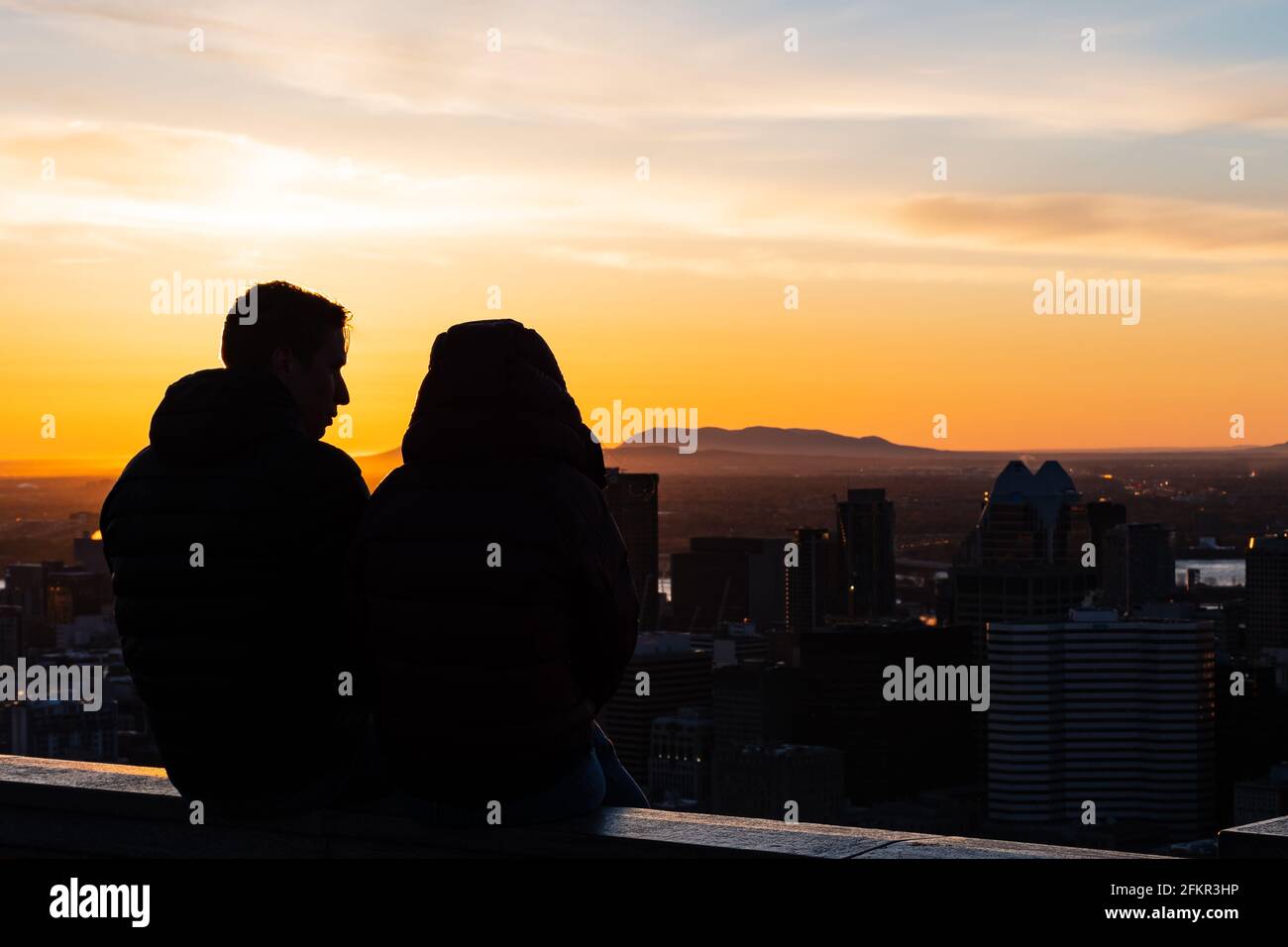 Back view of a couple sitting next to each other on the stone wall of the Kondiaronk belvedere and amiring the colorful view over Montreal's skyline Stock Photo