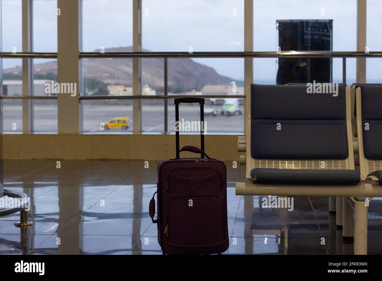 Unattended purple carry on suitcase by empty seat at airport terminal. Small lugagge left alone, security hazard concepts Stock Photo