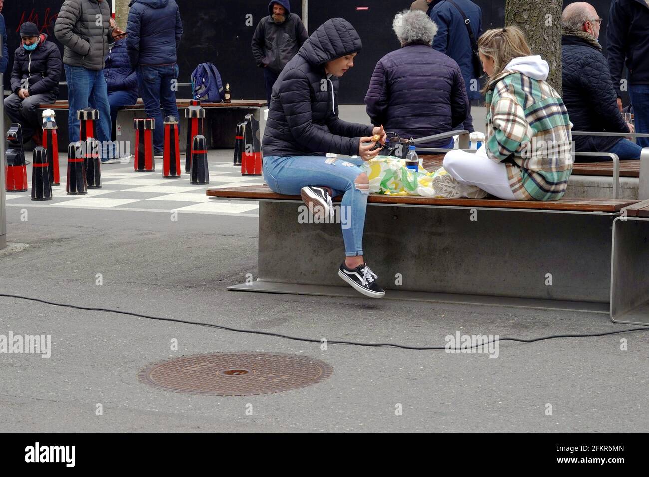 Street life in the city, in time of coronavirus and covid-19 crisis. People sit around, eat take away food, play chess and some of them wear face mask. Stock Photo