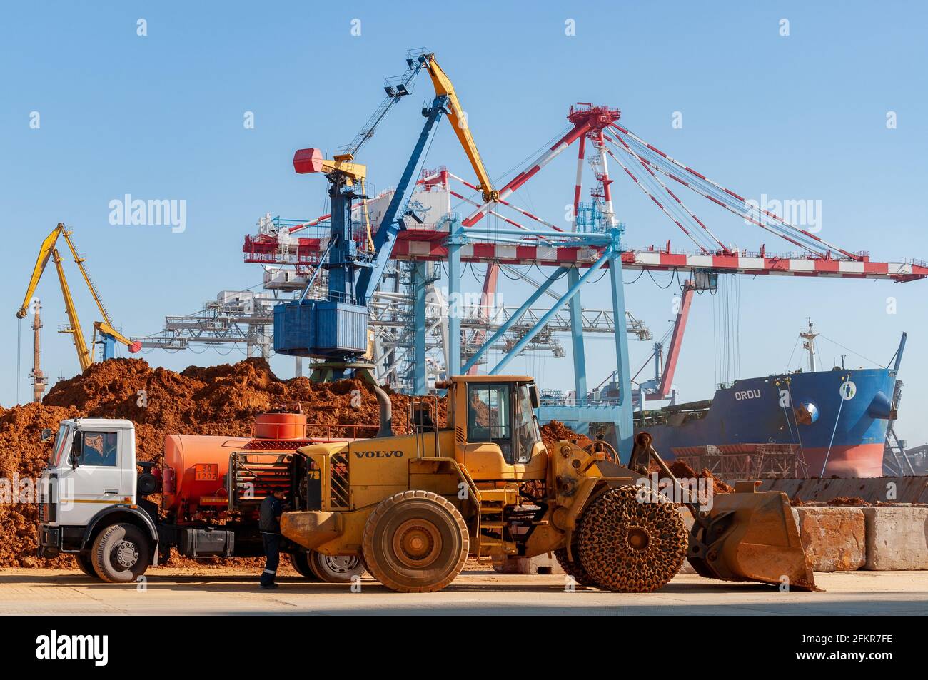 Container terminal of TIS Group dry cargo port. Construction truck and Volvo heavy bulldozer on port berth with moored cargo ship and shore cranes. Od Stock Photo