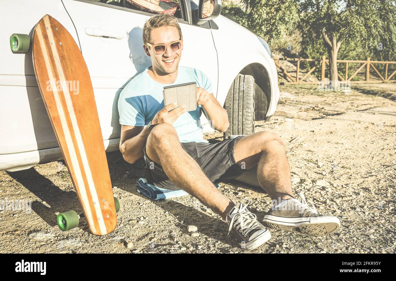 Young hipster fashion guy working remote on computer tablet sitting at car on road trip - New trend and technology concept with digital nomad Stock Photo