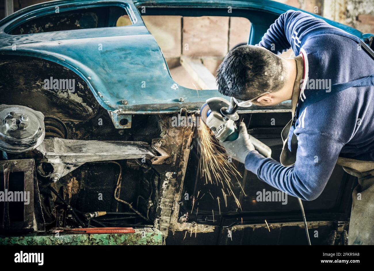 Young man mechanical worker repairing old vintage car body with electric grider in messy garage - Work safety with protection wear - Niche expertise Stock Photo