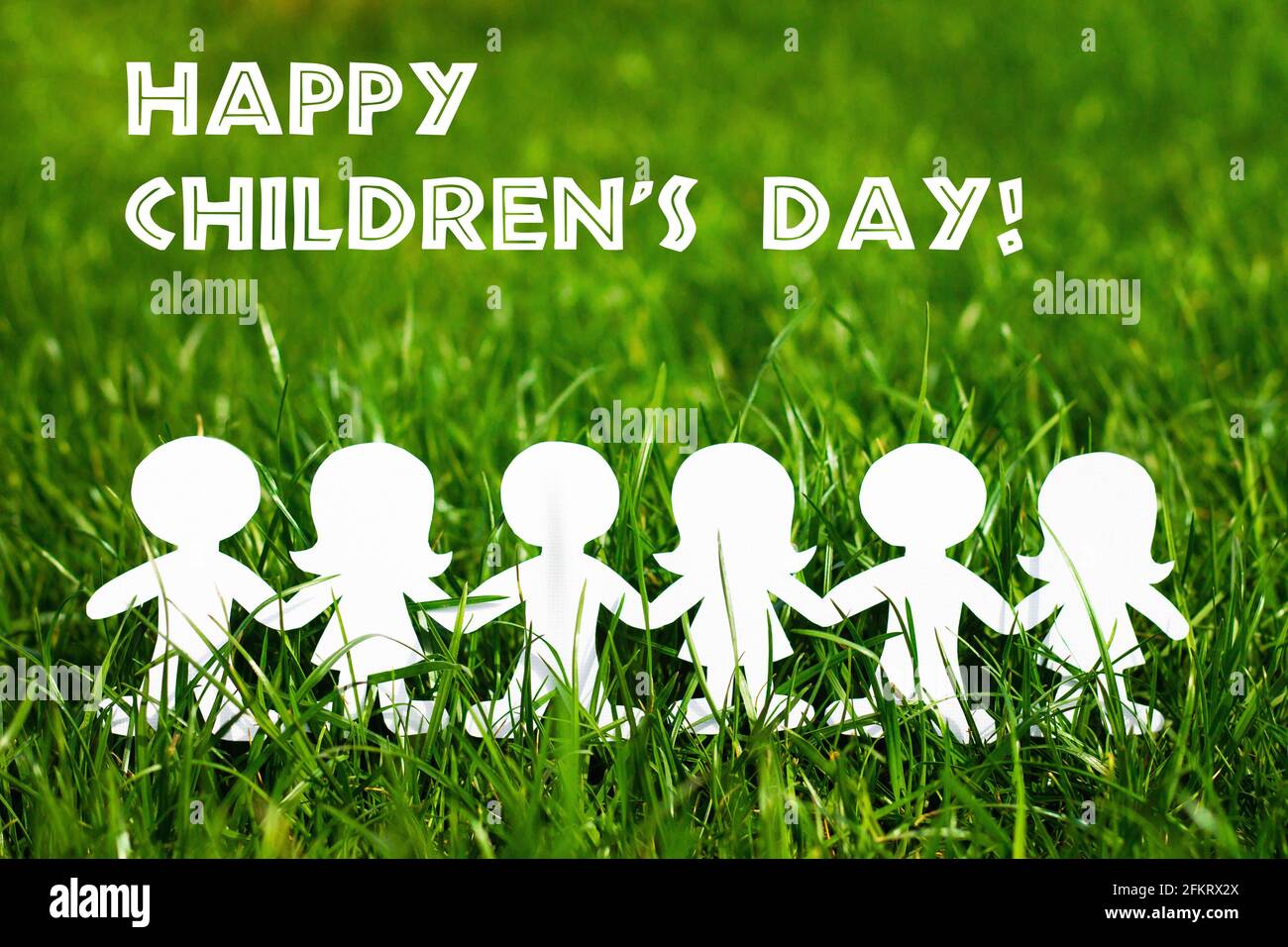 Silhouettes of children holding hands cut out of cardboard on a background of grass. Girls and boys made of white paper. International children's day. Stock Photo