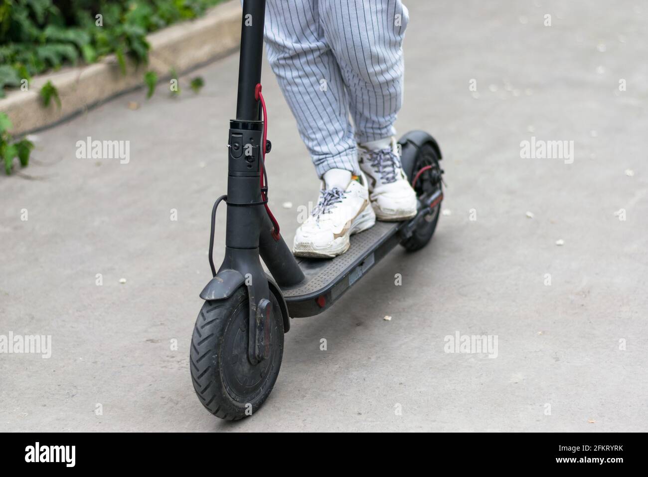 The legs of an unknown girl in white sneakers travel on a black electric scooter on a dark urban asphalt. Stock Photo