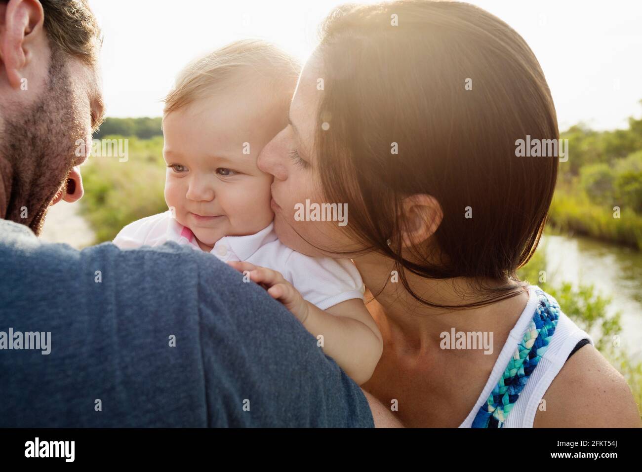 Over shoulder view of mid adult parents kissing baby daughter on riverside Stock Photo