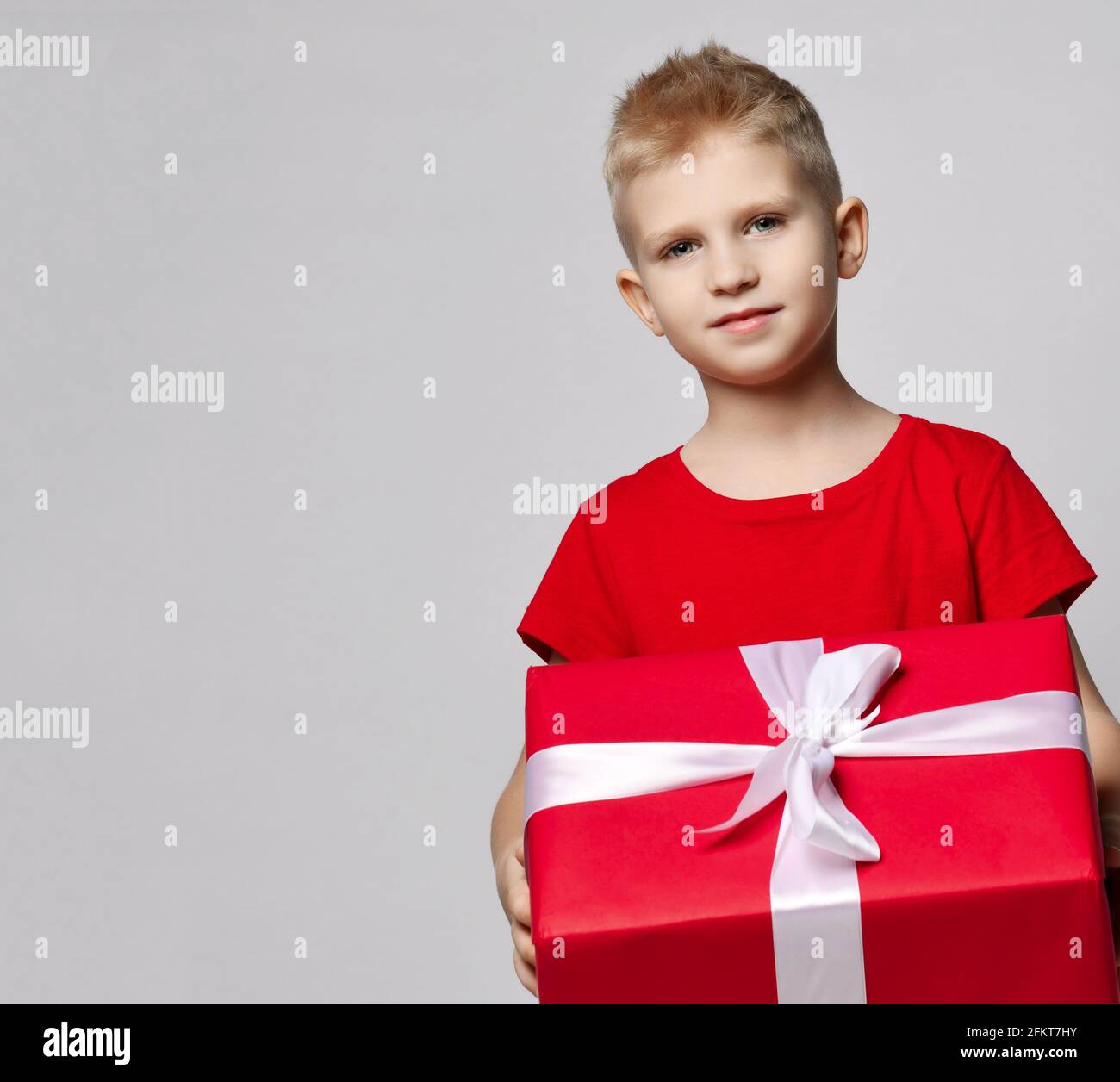 Portrait of smiling kid boy in red t-shirt holding big present box with ribbon in hands, carrying it, ready for birthday Stock Photo