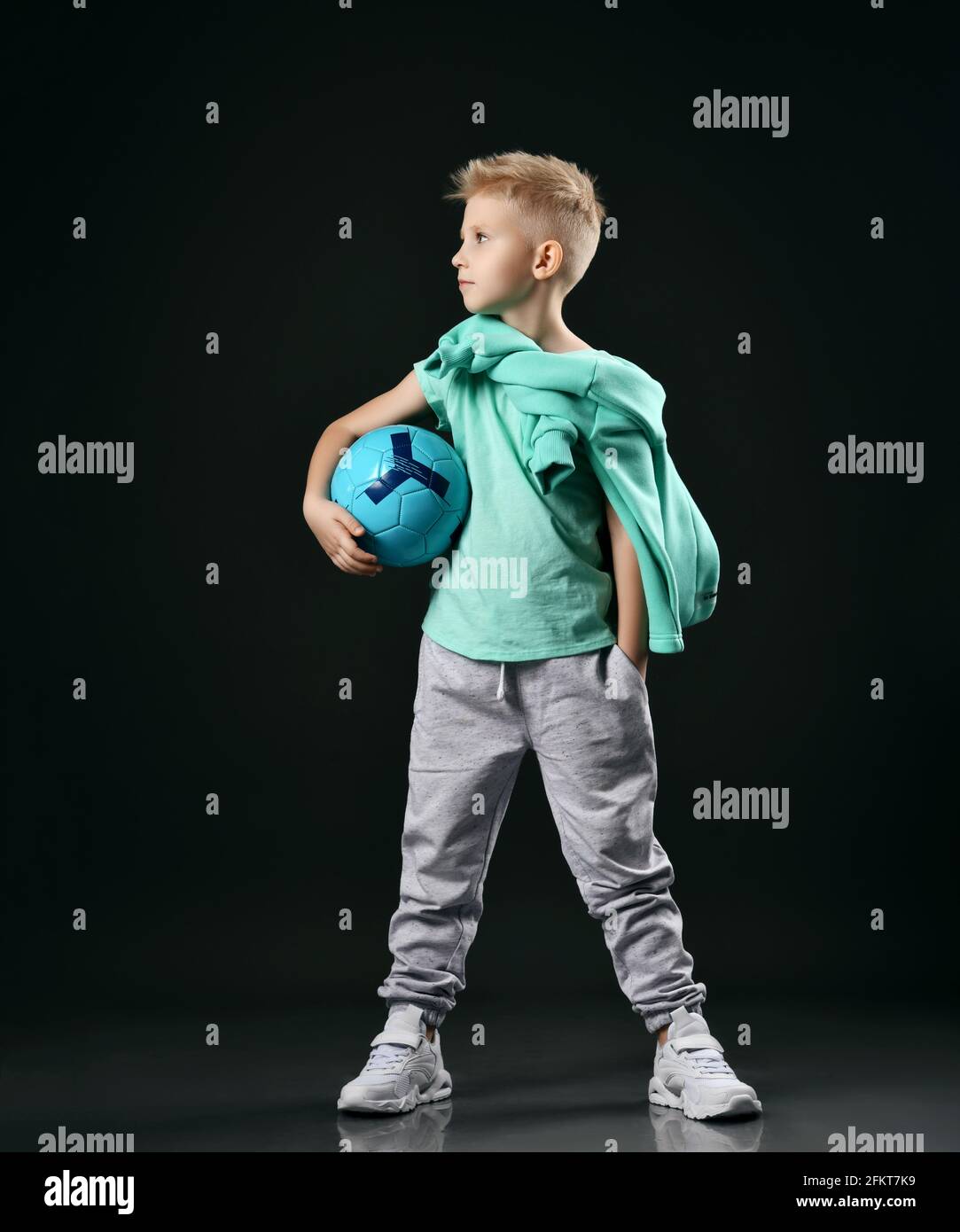 Schoolboy, teenager in casual t-shirt, pants, sneakers and hoodie around neck stands holding soccer ball in hand Stock Photo