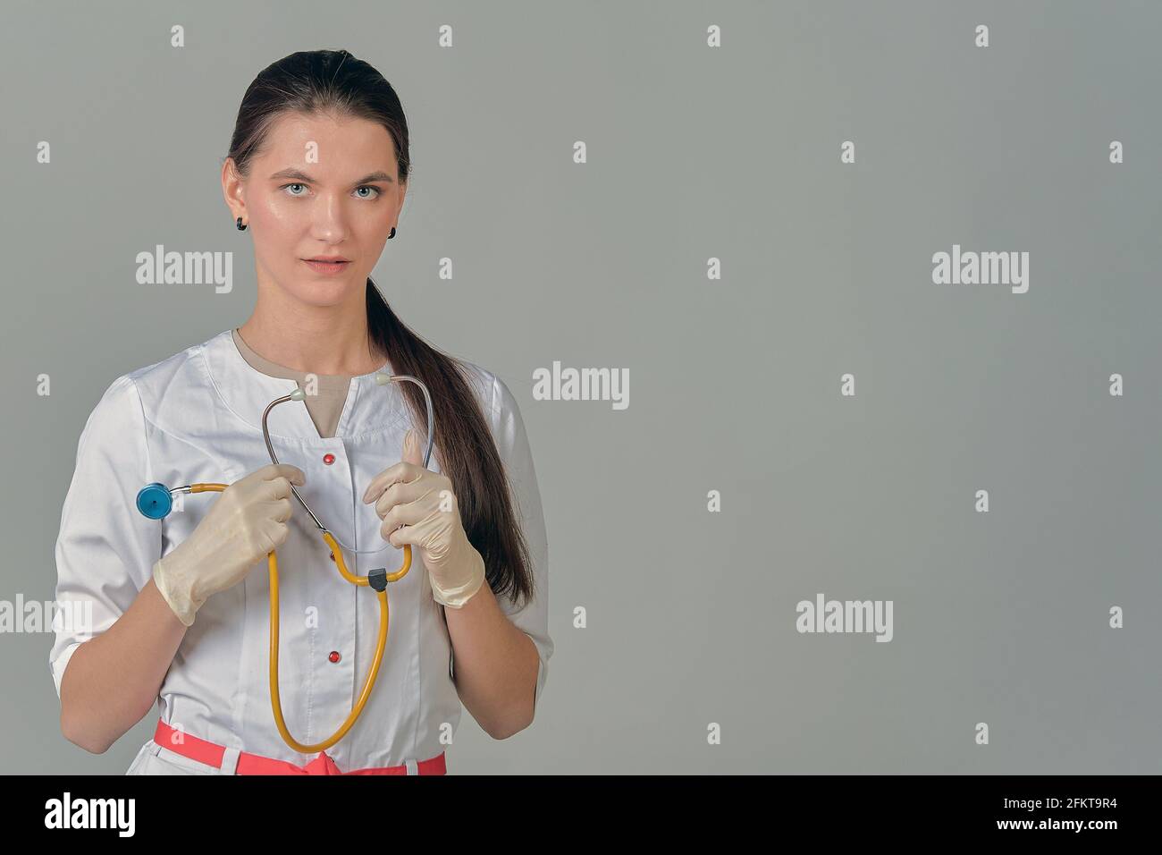 A young female doctor in a white lab coat with a stethoscope in her hands on a grey background Stock Photo