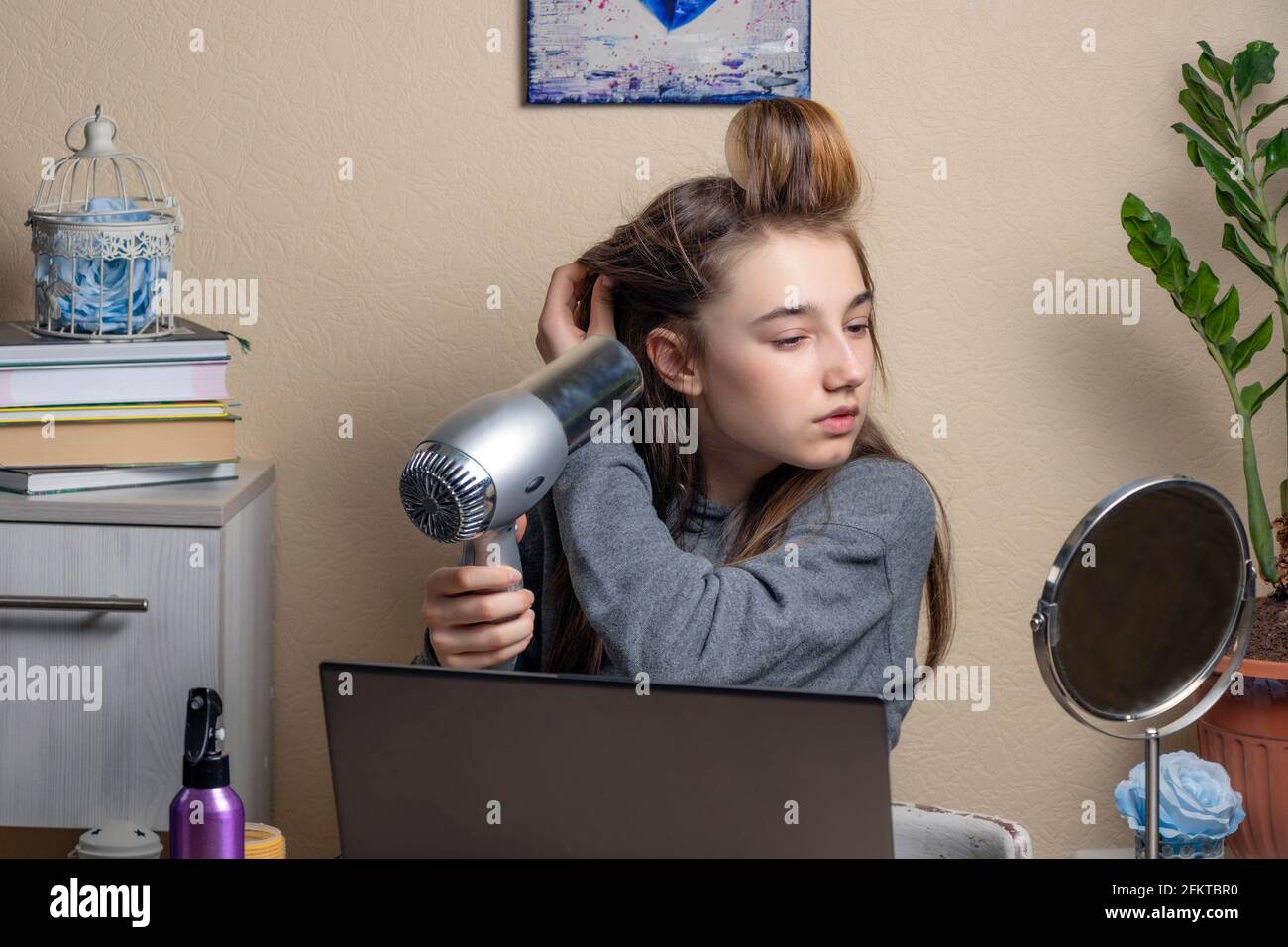 teenage girl with curlers and a hairdryer looks at the laptop screen. beautiful teenage girl doing her hair in front of laptops. Online beauty concept Stock Photo