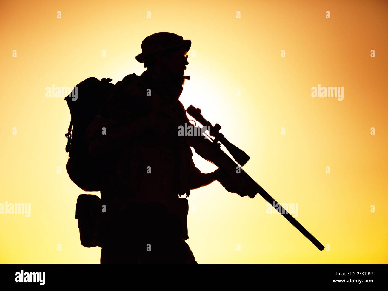 Silhouette of army elite forces sniper, commando shooter in boonie hat, carrying backpack, equipped tactical radio, walking on background of setting Stock Photo