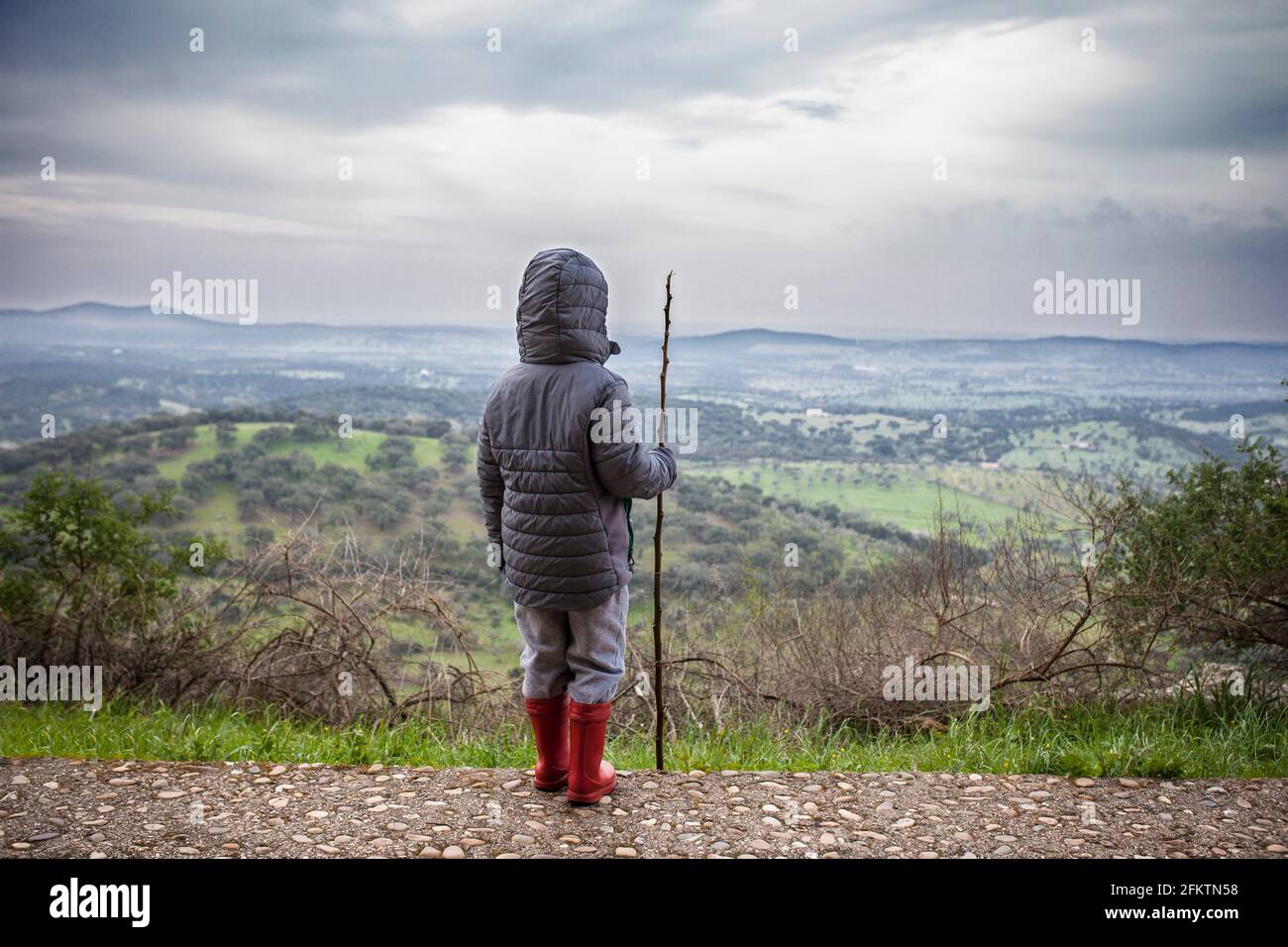 Child boy observing the dehesa landscape outskirts Alburquerque, Extremadura, Spain. He holds a cane over cloudy sky. Stock Photo
