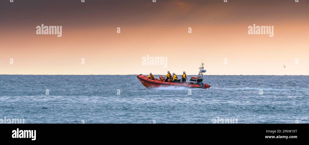 A panoramic image of RNLI Gladys Mildred, Newquay’s Atlantic 85 lifeboat responding to an emergency callout at the end of the day in Fistral Bay in Co Stock Photo