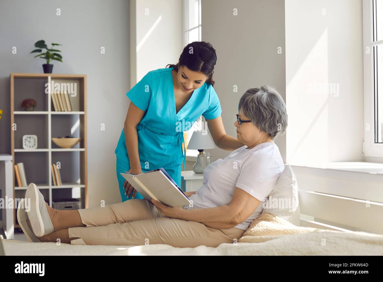 Home care nurse talking to relaxed senior woman who's sitting on bed and reading book Stock Photo