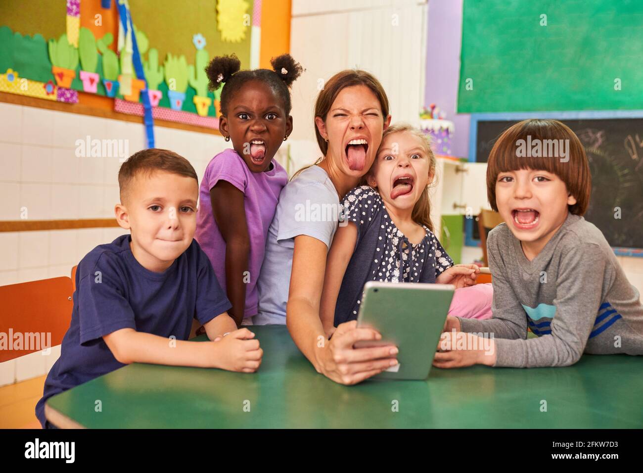 Children and kindergarten teacher with tablet computer stick their tongue out in kindergarten Stock Photo