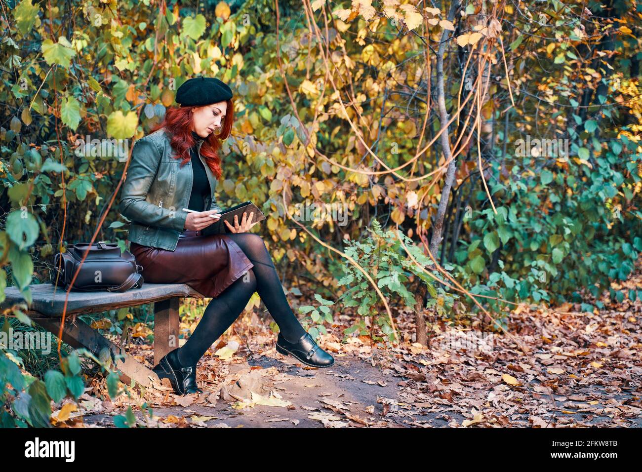 Woman with graphic tablet on park bench. Autumn landscape in the background. Red-haired girl in a beret and a fashionable jacket with a leather backpack. Designer or freelancer in nature. Stock Photo