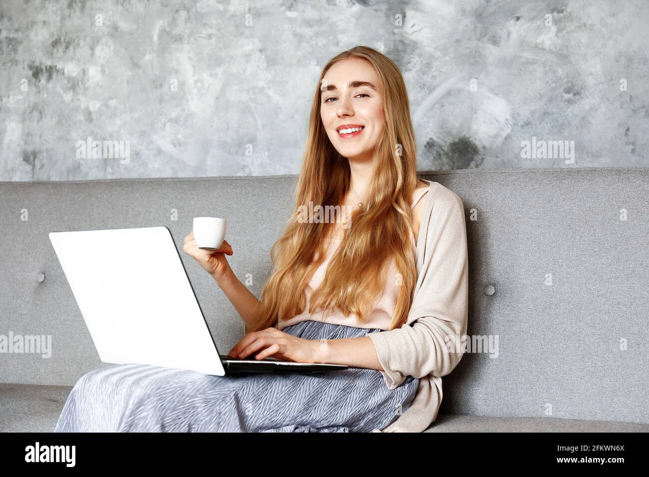 Young beautiful woman wearing white t-shirt on grey textile sofa at home. Attractive slim female in domestic situation, resting on couch in her lofty Stock Photo