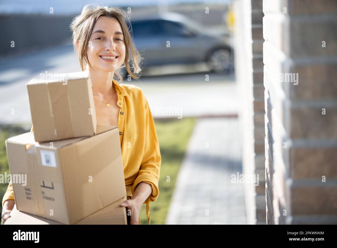 Young happy woman carries home a parcels with goods purchased online, knocking on the door. Concept of online shopping and delivery. Looking at camera. Stock Photo