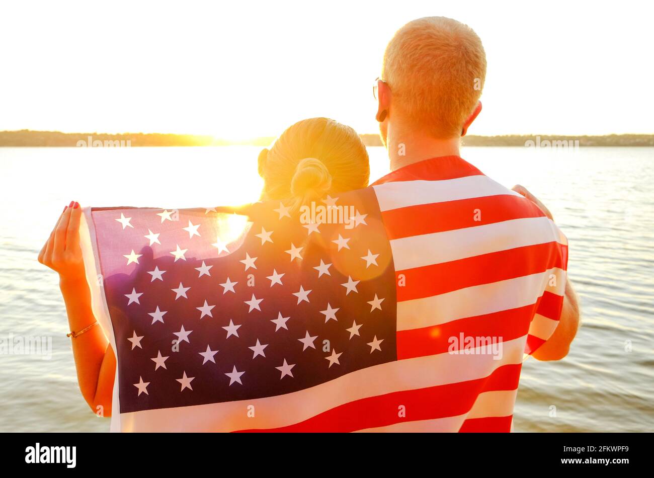 Rear view of young American patriotic couple hugging with USA flag on their backs enjoying beautiful summer sunset by the river. Independence day cele Stock Photo