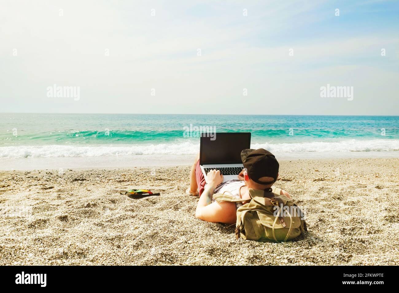 Back view, hipster young man lying on sandy beach leaning on backpack, with laptop computer, typing, blogging, browsing. Freelance, distance work conc Stock Photo