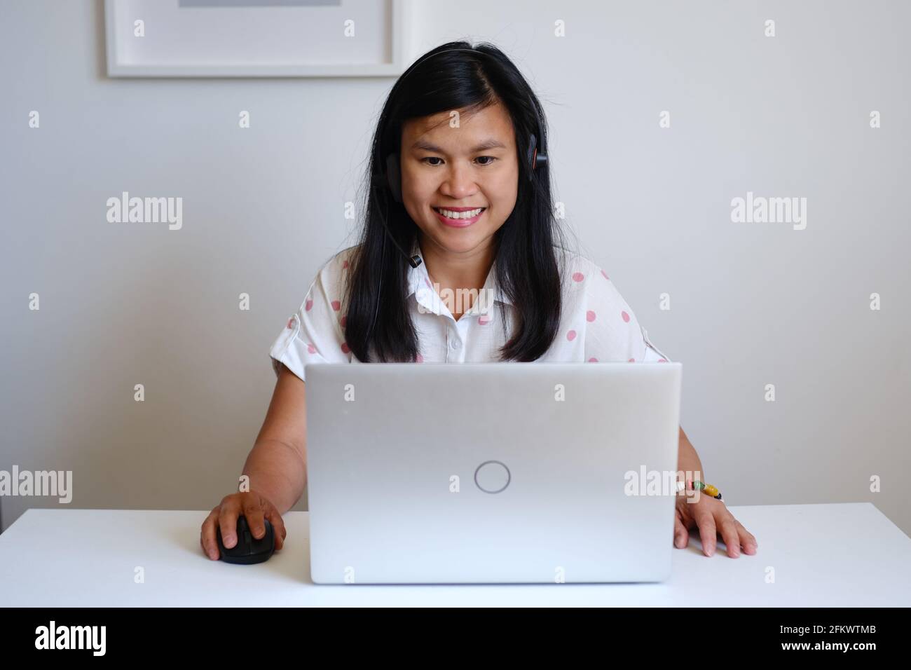 Filipino filipina Asian woman or girl teacher working sitting in front of a white desk and silver computer, teaching a lesson online on the Internet Stock Photo