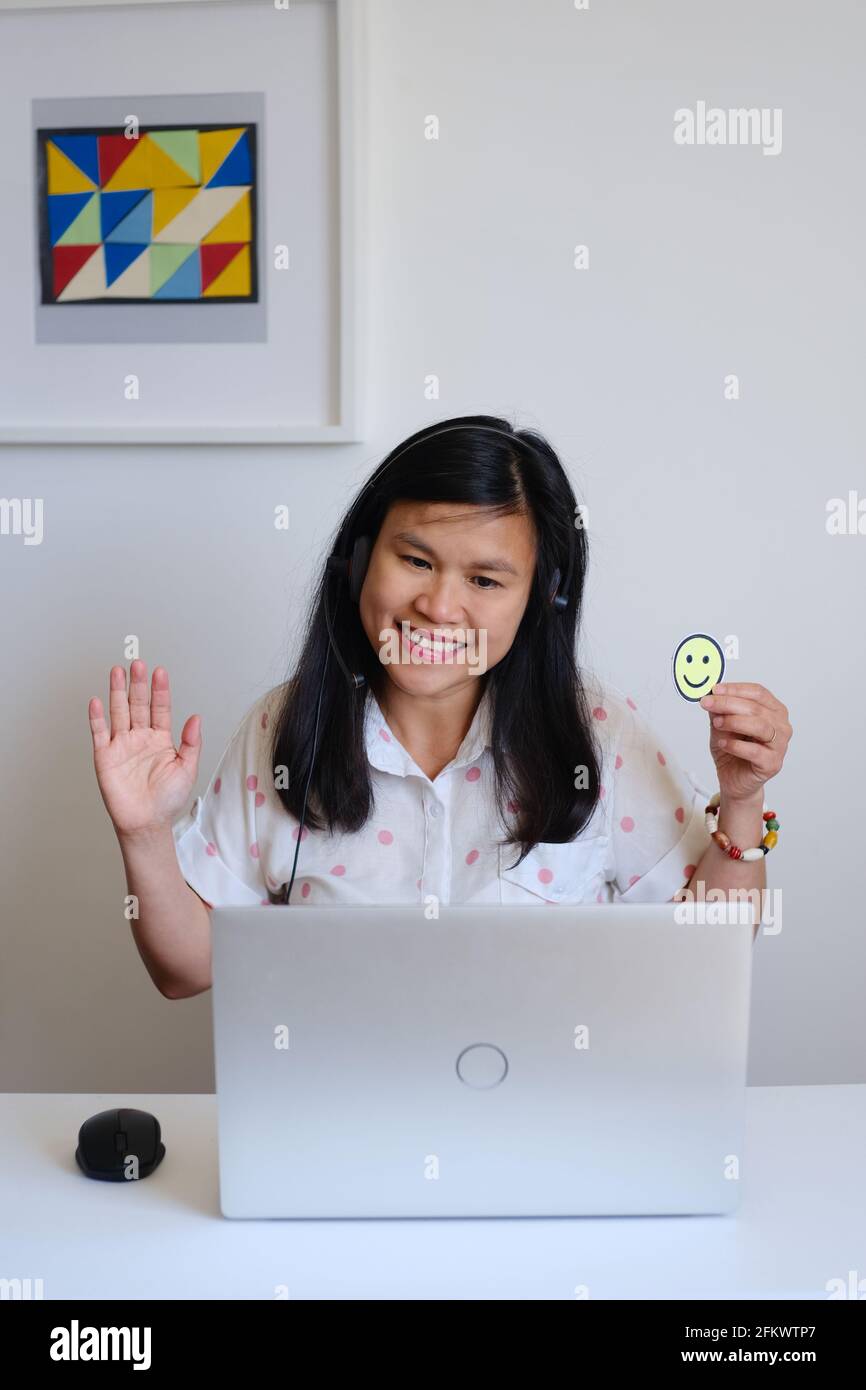 Filipino filipina Asian woman or girl teacher working sitting in front of a white desk and silver computer, teaching a lesson online on the Internet Stock Photo