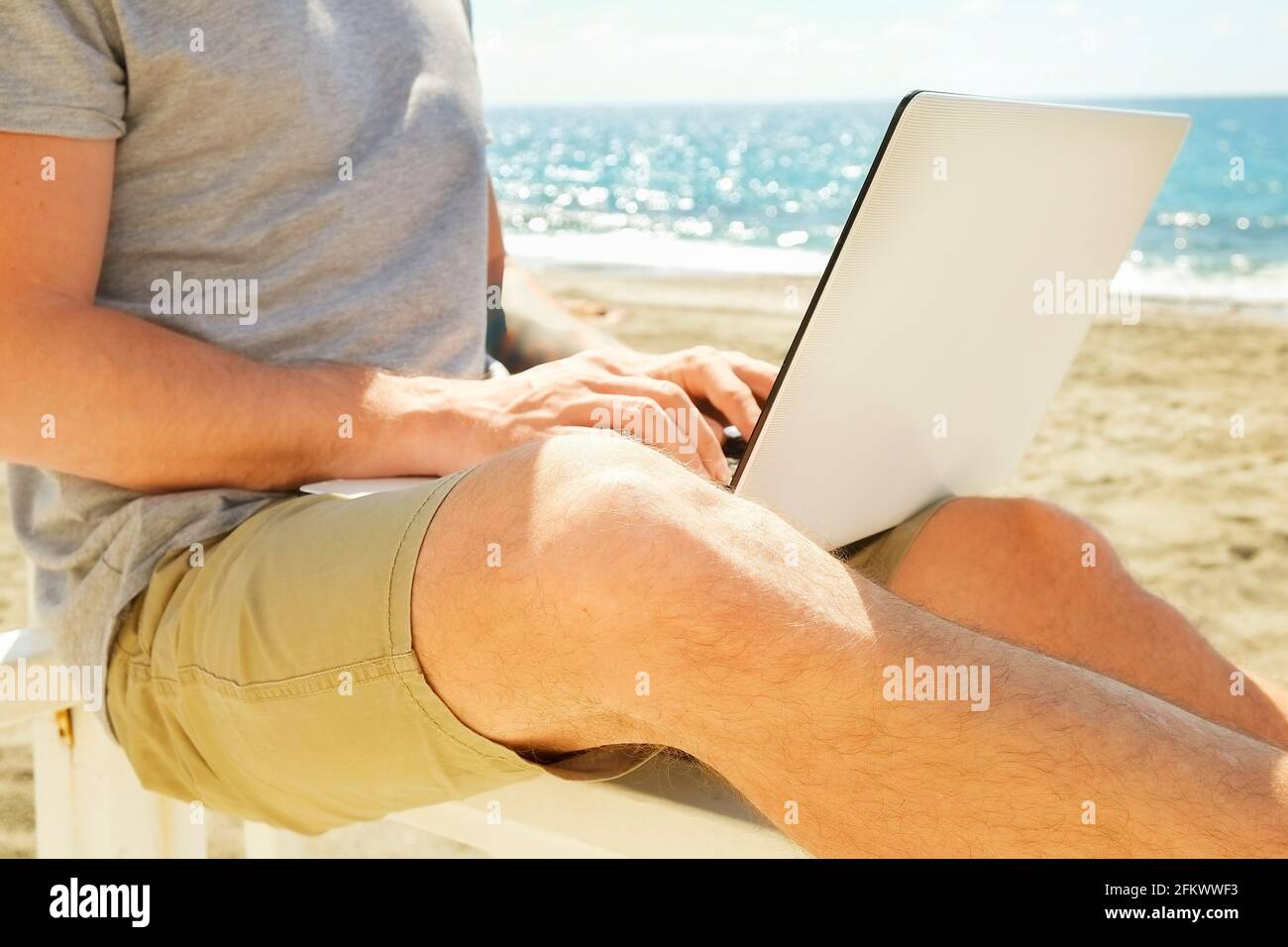 Fit young man using laptop sitting on white wood beach veranda porch. Perks of freelance concept. Male programmer working coding on notebook computer. Stock Photo