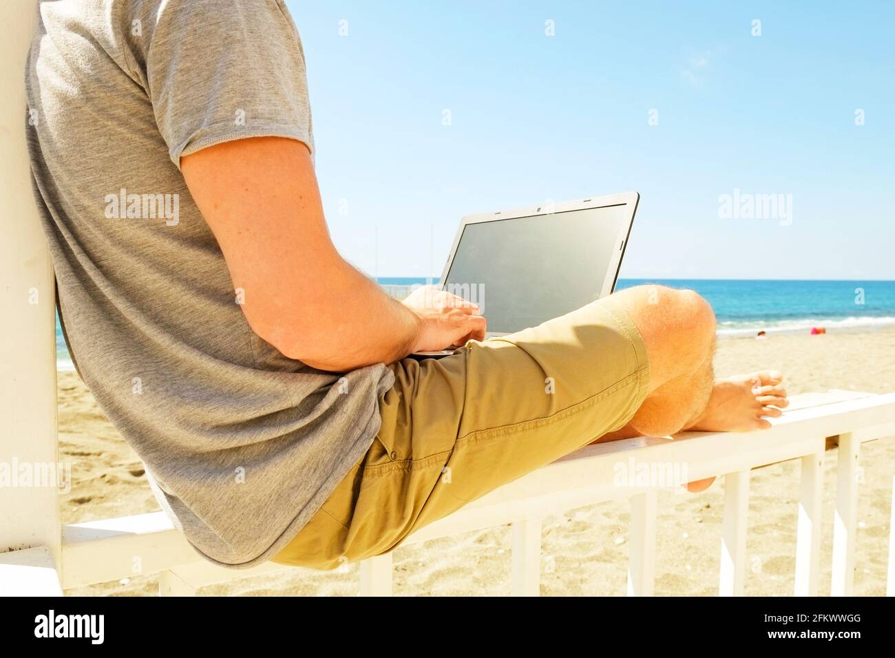 Fit young man using laptop sitting on white wood beach veranda porch. Perks of freelance concept. Male programmer working coding on notebook computer, Stock Photo