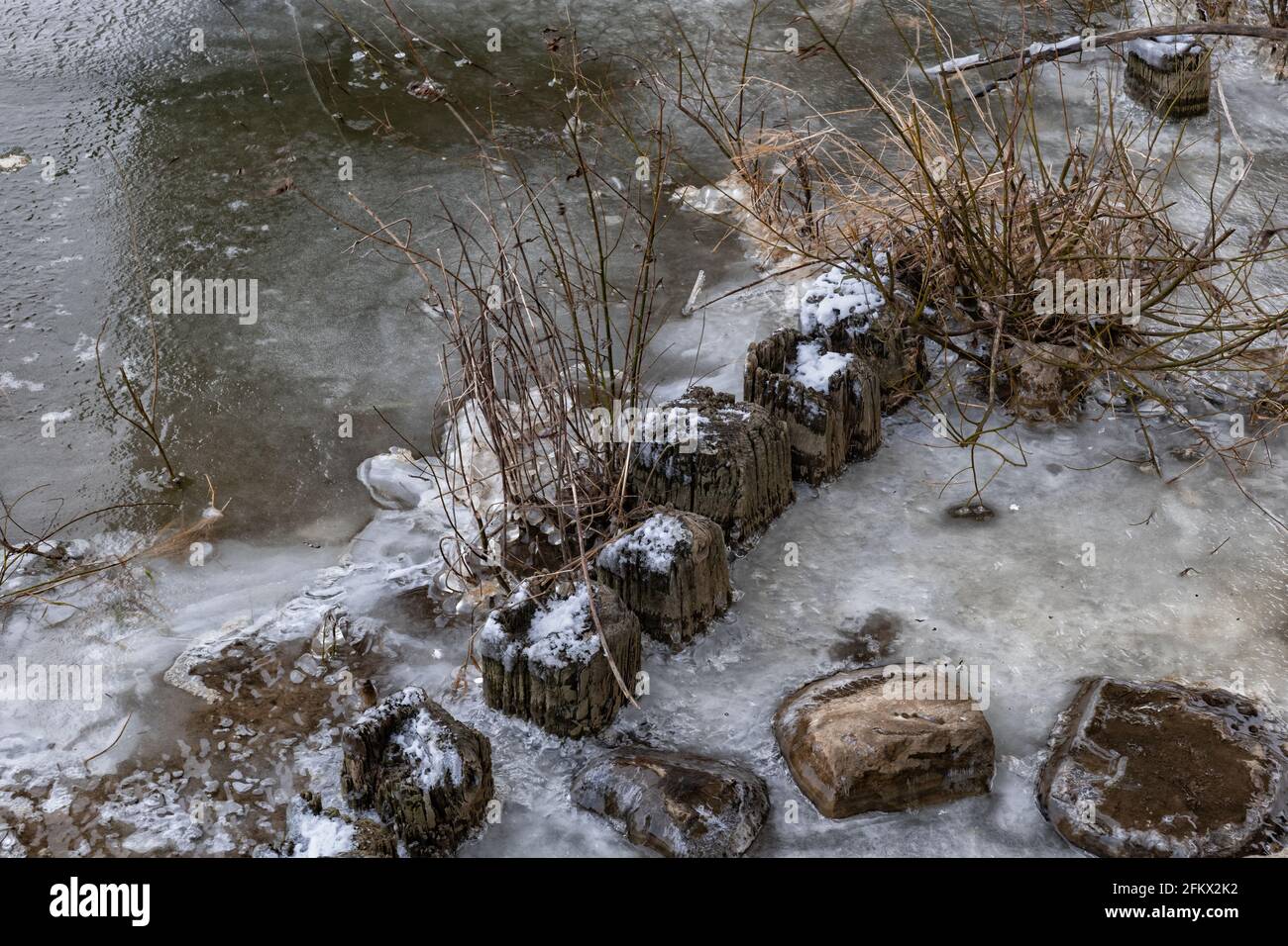 Frozen river with old weathered wooden posts and rocks, winter season abstract background. Stock Photo