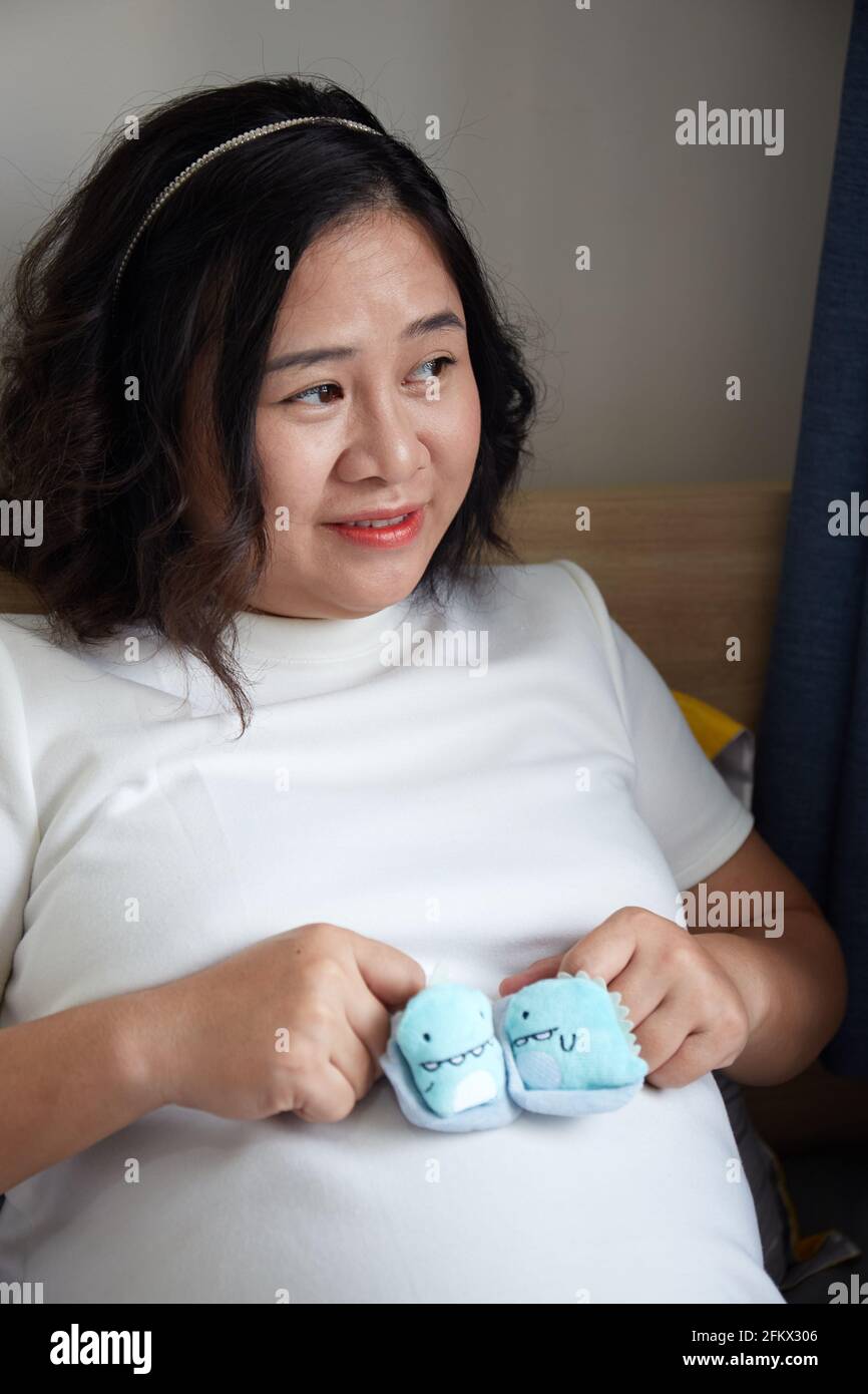 Young Vietnamese pregnant woman with short hair holding baby shoes on