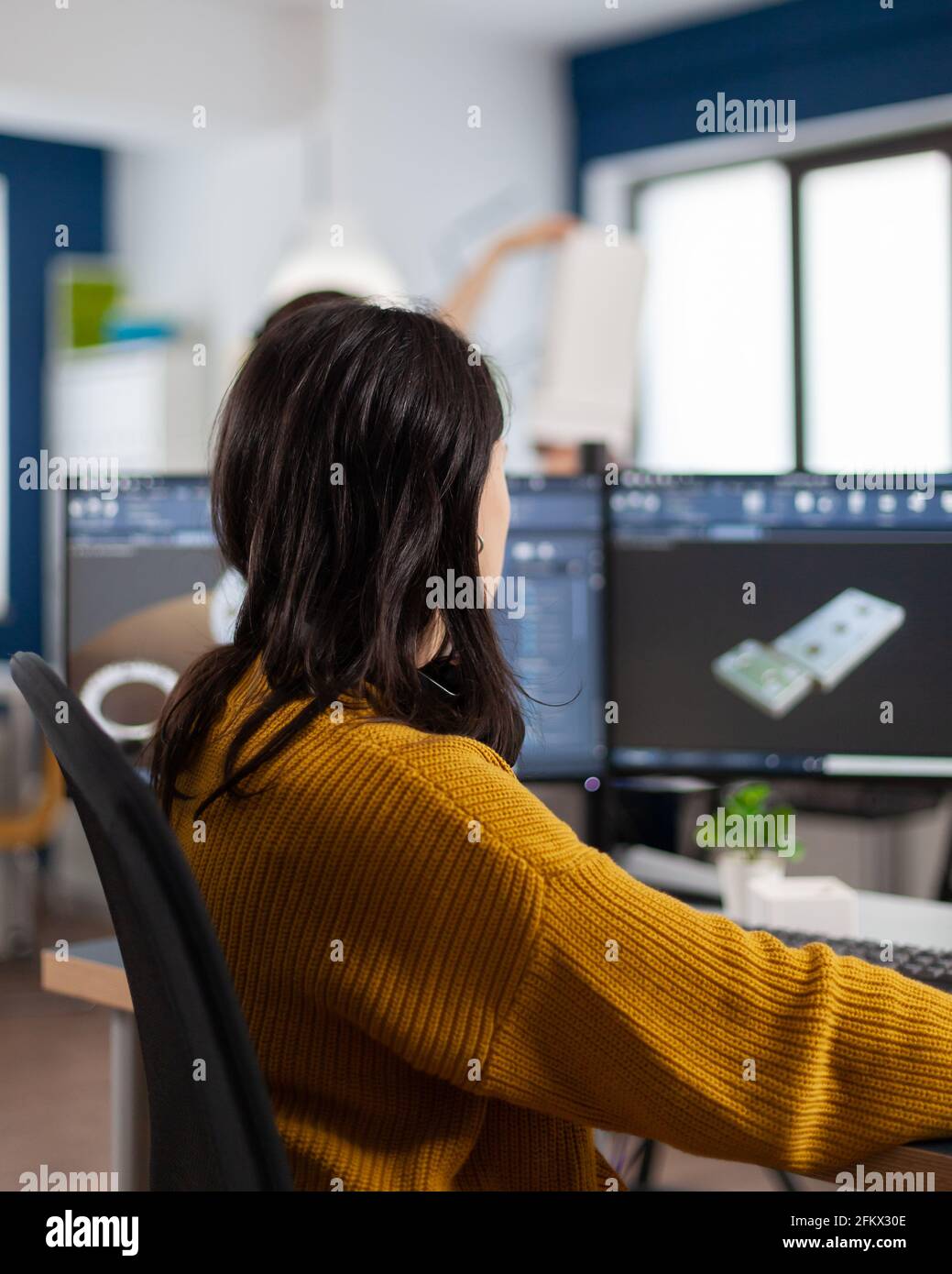 Young woman industrial engineer working at pc with two monitors, screens showing CAD software with 3D prototype of gears metalic mechanical piece. Employee working in creative agency office Stock Photo