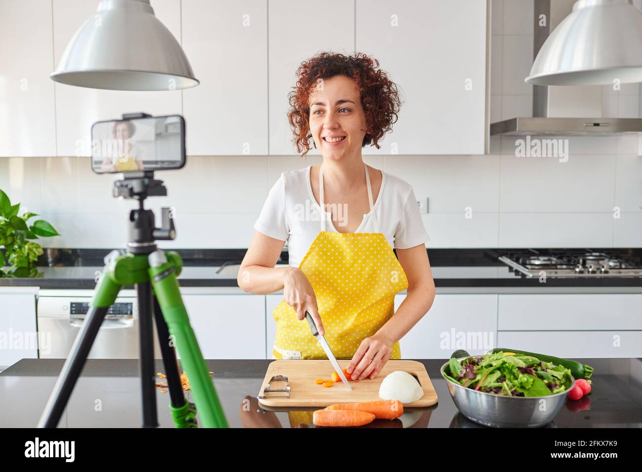 Woman youtuber records herself with her smartphone in her kitchen while preparing a salad recipe, there is natural light and she is wearing a yellow a Stock Photo