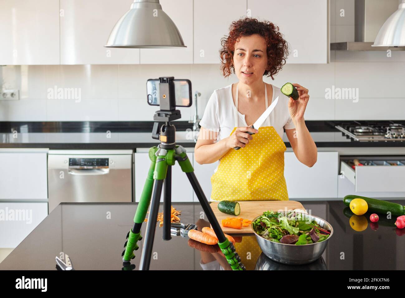 Woman youtuber records herself with her smartphone in her kitchen while preparing a salad recipe, there is natural light and she is wearing a yellow a Stock Photo