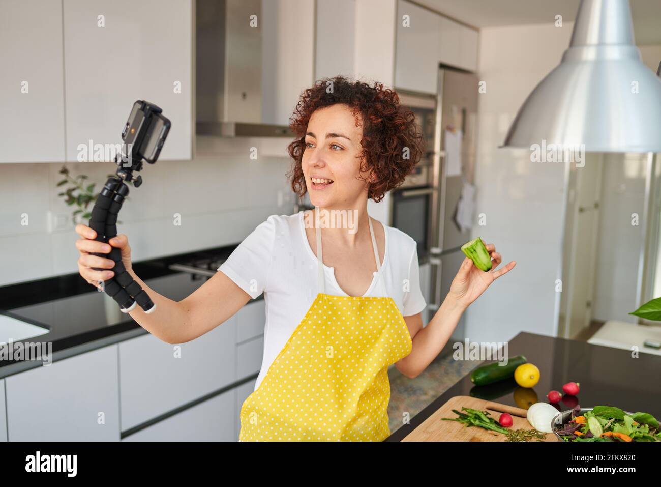 Woman youtuber records herself with her smartphone in her kitchen while preparing a salad recipe, there is natural light and she is wearing a yellow a Stock Photo