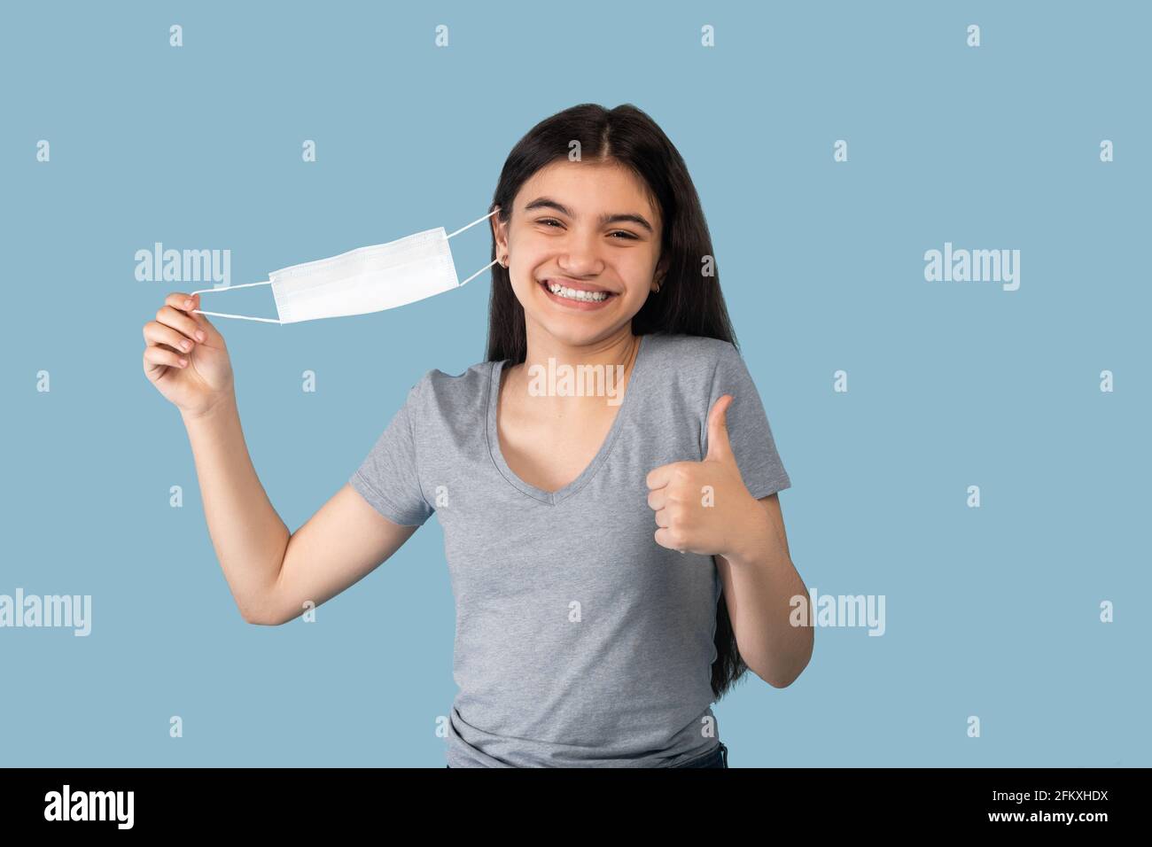 Coronavirus is over. Happy Indian teenage girl taking off her face mask and showing thumb up gesture on blue background Stock Photo
