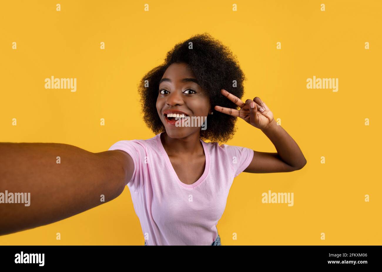Positive african american woman taking selfie and showing peace gesture, smiling at camera over yellow background Stock Photo