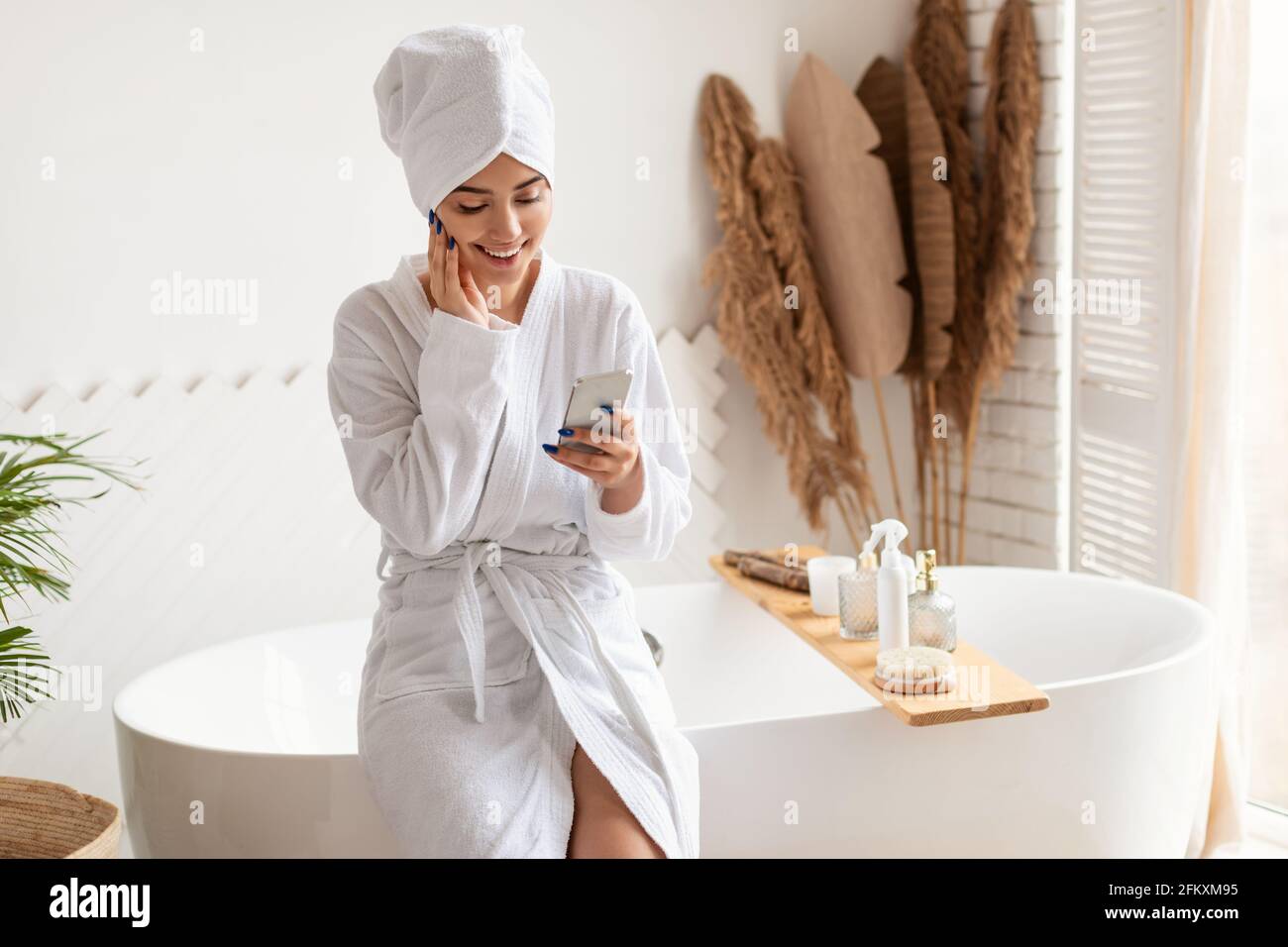 Woman Using Smartphone In Bathroom Wearing Bathrobe Sitting On Bathtub Stock Photo