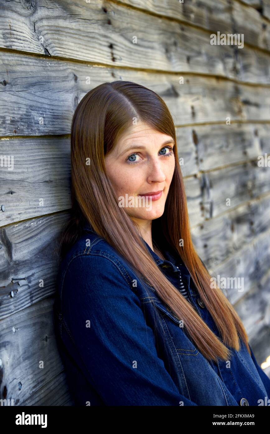 Young woman leans against a rustic wooden wall and looks doubtfully at the camera.  She is wearing a denim jacket and has long brown hair. Stock Photo