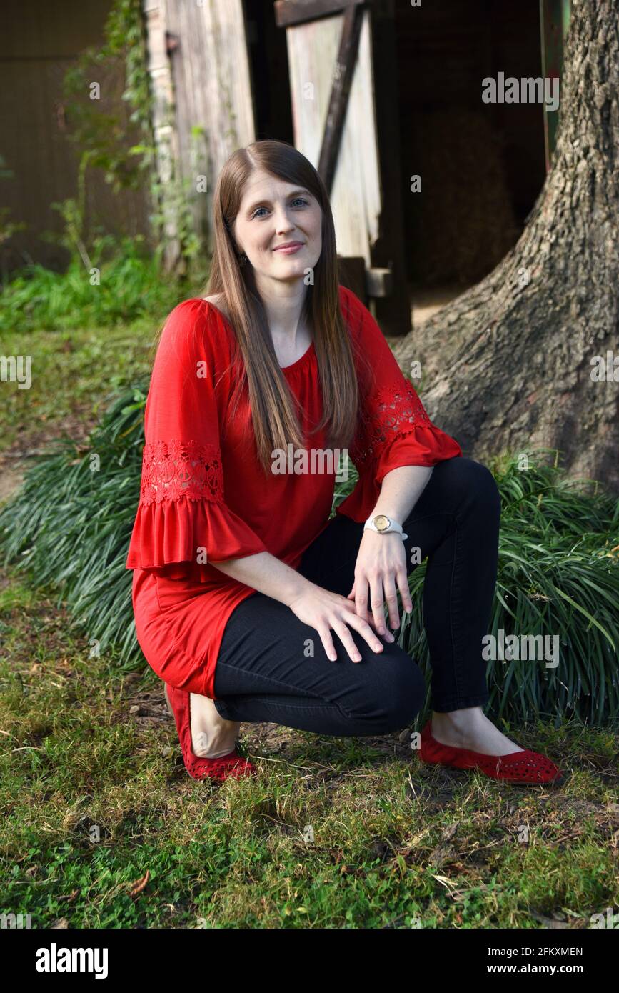 Young woman kneels outside in front of a tree and rustic, wooden barn.  She is unsmiling and is wearing red blouse and red shoes. Stock Photo