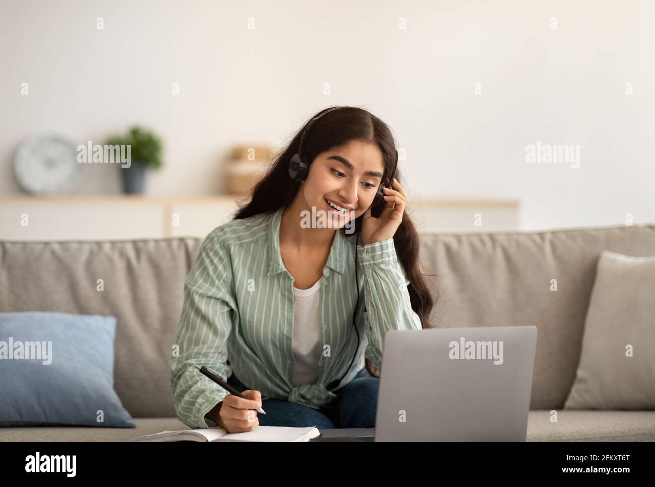 Happy Indian businesswoman in headphones having online business meeting on laptop, writing down info at home Stock Photo