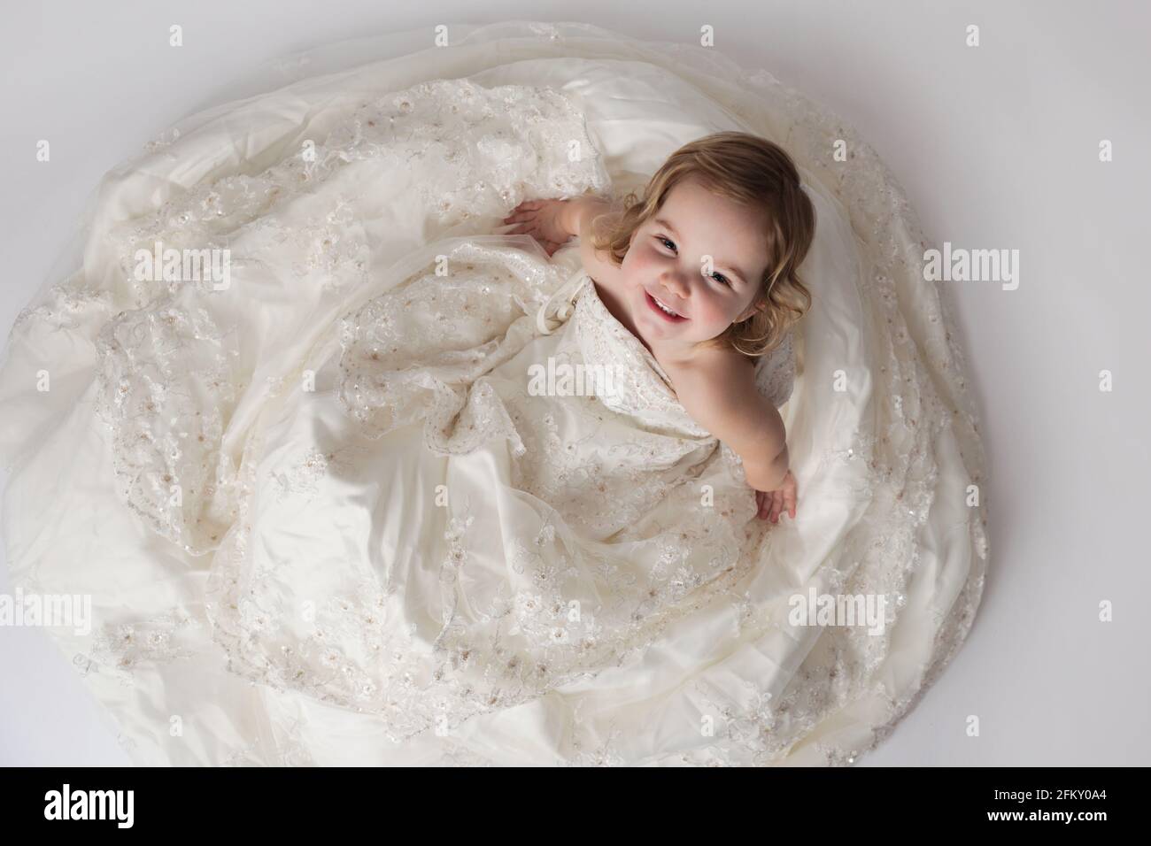 Toddler girl looks up and smiles while wearing mother's wedding gown Stock Photo