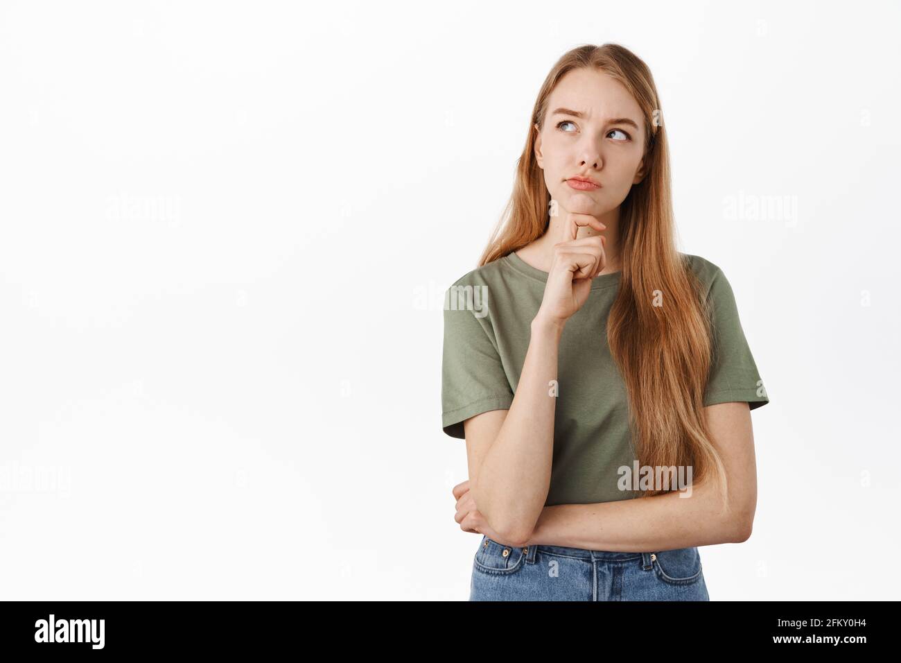 Thoughtful young woman making serious decision, looking up while pondering choices, frowning while thinking, standing in t-shirt and jeans against Stock Photo