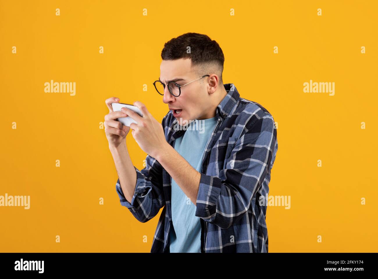 Online gaming. Addicted young guy playing video games on cellphone, overusing his gadget on orange studio background Stock Photo